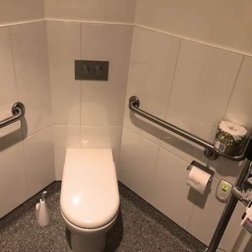 A white ceramic toilet in a compact, accessible hospital bathroom with white tiled walls, stainless steel grab handles, a wall-mounted toilet paper roll dispenser, and a speckled gray linoleum floor.