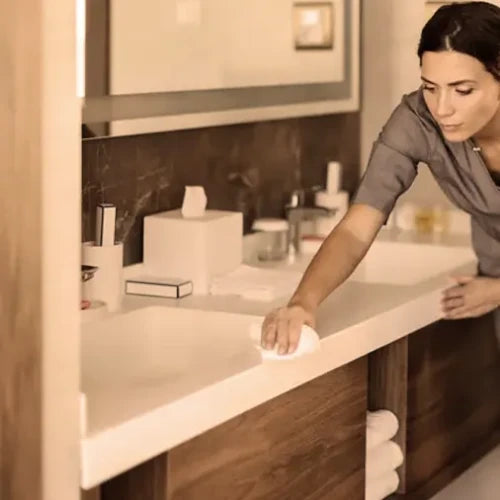 A woman in a gray top wipes down a white bathroom countertop with a white cloth, with a sink, mirror, and wooden vanity in the background. In a hotel room.